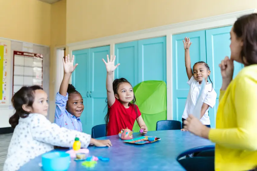 A group of children sitting at a table with their hands raised.