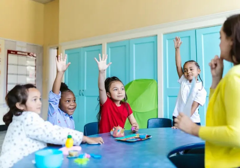 A group of children sitting at a table with their hands raised.